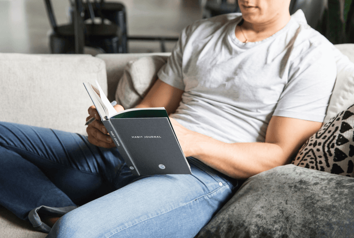 Man in White T-shirt and Jeans Sitting on a Couch Flipping Through Habit Journal